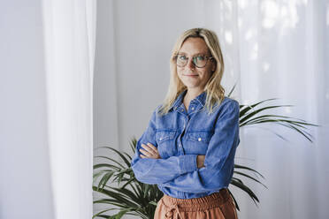 Young woman wearing eyeglasses standing with arms crossed at home - EBBF05773