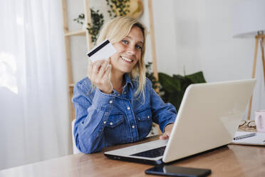 Smiling freelancer with laptop showing credit card at home office - EBBF05785