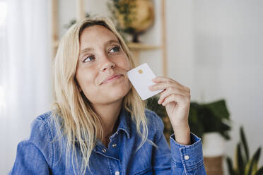 Thoughtful blond woman holding credit card at home - EBBF05788