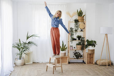 Young woman with arms outstretched standing on stool at home - EBBF05802