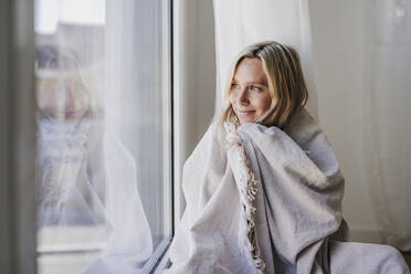 Junge Frau in Decke eingewickelt, die zu Hause aus dem Fenster schaut - EBBF05806