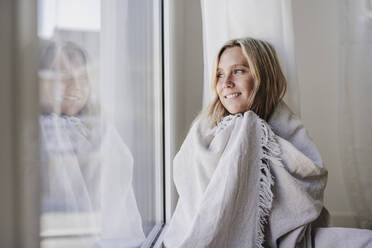 Lächelnde Frau, die zu Hause in eine Decke eingewickelt aus dem Fenster schaut - EBBF05807