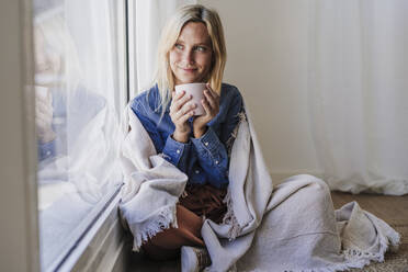 Young woman wrapped in blanket holding coffee cup sitting by window at home - EBBF05809