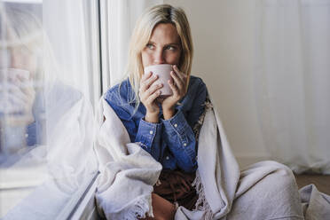 Woman drinking coffee sitting by window wrapped in blanket at home - EBBF05810