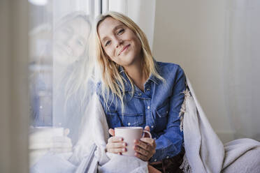 Lächelnde Frau mit Kaffeetasse, die sich an ein Glasfenster zu Hause lehnt - EBBF05812