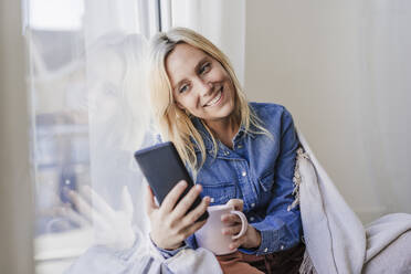 Lächelnde Frau, die ein Smartphone benutzt und sich zu Hause an eine Fensterscheibe lehnt - EBBF05813