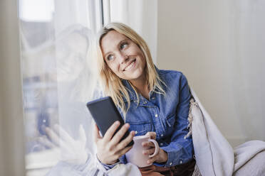 Frau mit Smartphone schaut aus dem Fenster und sitzt zu Hause - EBBF05814