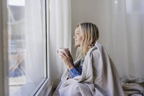 Lächelnde, in eine Decke eingewickelte Frau mit einer Tasse in der Hand, die zu Hause aus dem Fenster schaut - EBBF05815