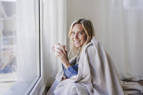 Junge Frau, in eine Decke eingewickelt, sitzt am Fenster und hält eine Tasse zu Hause - EBBF05816