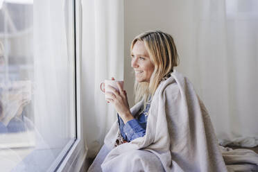 Lächelnde blonde Frau mit Kaffeetasse, die zu Hause aus dem Fenster schaut - EBBF05817