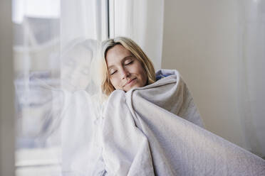 Woman with eyes closed wrapped in blanket relaxing by window at home - EBBF05819