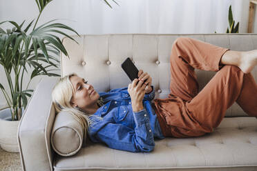 Frau mit Smartphone auf dem Sofa liegend zu Hause - EBBF05822