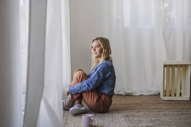 Woman sitting on carpet hugging knees near window at home - EBBF05825