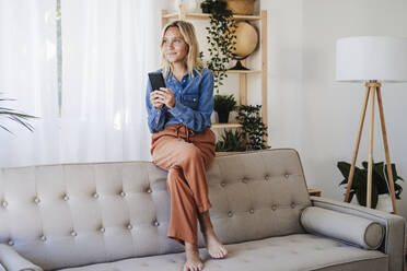 Lächelnde Frau mit Smartphone sitzt zu Hause auf dem Sofa - EBBF05826