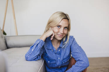 Lächelnde blonde junge Frau, die zu Hause auf dem Sofa sitzt - EBBF05827