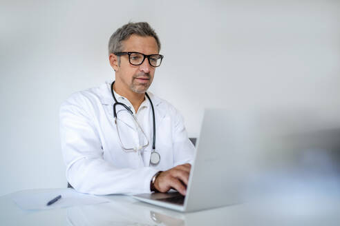 Älterer Arzt mit Laptop am Schreibtisch in einer Arztpraxis - DIGF18570