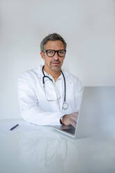 Porträt eines reifen Arztes mit Laptop am Schreibtisch in einer Arztpraxis - DIGF18571