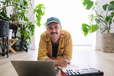 Happy man with laptop and piano lying on floor at home - ASGF02605