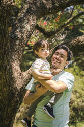 Glücklicher Vater mit Sohn vor einem Baum im Park stehend - FLMF00844