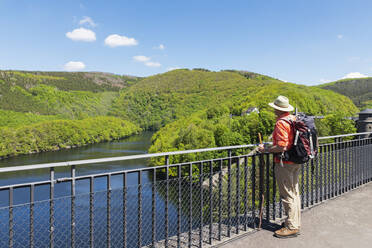 Senior man wearing hat and backpack standing by railing looking at view, Eifel National Park, Germany - GWF07529