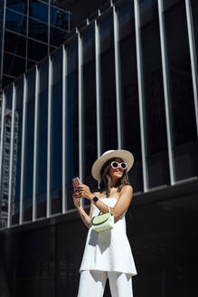 Fashionable young woman with smart phone standing outside building - DAMF01070