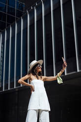 Junge stilvolle Frau nimmt Selfie durch Smartphone außerhalb des Gebäudes an einem sonnigen Tag - DAMF01071
