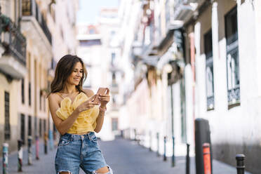 Happy young woman using mobile phone amidst buildings on sunny day - DAMF01080