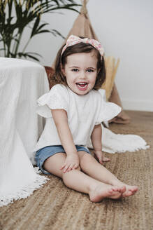 Glückliches süßes Mädchen sitzt auf Teppich im Wohnzimmer - EBBF05847