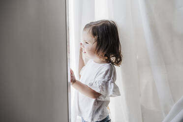 Cute little girl looking out of window at home - EBBF05849