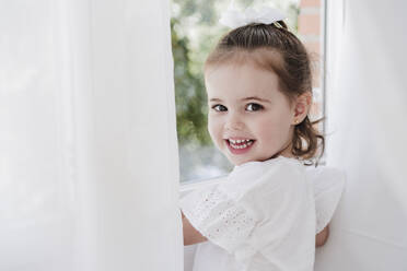 Happy cute girl standing by window at home - EBBF05867