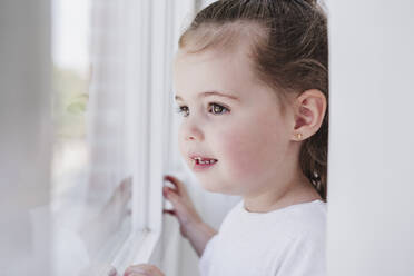 Nachdenkliches süßes Mädchen, das zu Hause aus dem Fenster schaut - EBBF05868
