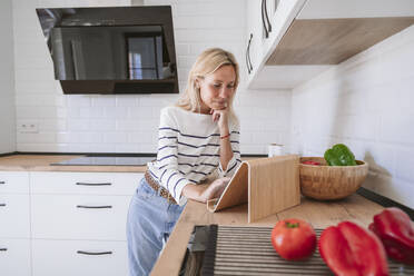 Young woman with hand on chin watching recipe on tablet PC in kitchen at home - EBBF05886