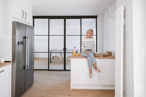 Young woman sitting on counter in domestic kitchen at home - EBBF05896