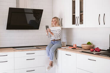 Lächelnde Frau hält Handy auf Küchentisch zu Hause - EBBF05912