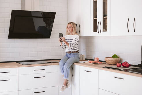 Smiling woman holding mobile phone on kitchen counter at home - EBBF05912