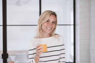 Smiling woman holding orange juice glass at home - EBBF05918