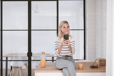 Thoughtful woman with smart phone on kitchen counter at home - EBBF05920