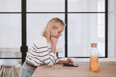 Young woman using mobile phone on kitchen counter at home - EBBF05922