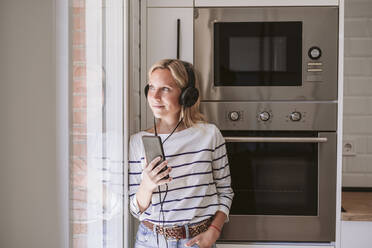 Frau mit Mobiltelefon, die am Fenster zu Hause Musik über Kopfhörer hört - EBBF05924
