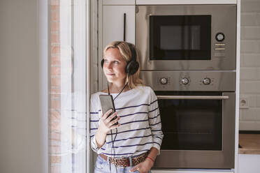 Woman with mobile phone listening music on headphones by window at home - EBBF05924