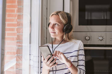 Junge Frau hört Musik über Kopfhörer am Fenster zu Hause - EBBF05925
