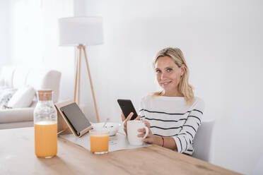 Lächelnde Frau mit Kaffeetasse und Mobiltelefon zu Hause - EBBF05945