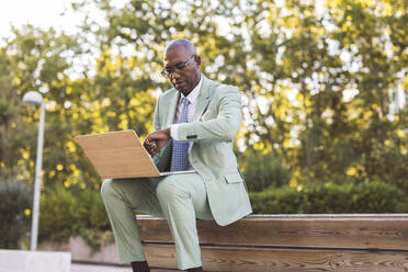 Älterer Geschäftsmann mit Laptop, der die Zeit im Park kontrolliert - JCCMF07057