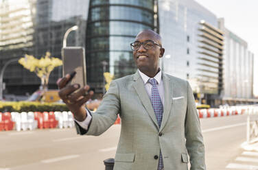 Smiling mature businessman taking selfie through smart phone on street - JCCMF07076