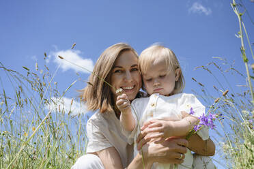 Happy mother with daughter sitting under sky at field - EYAF02025