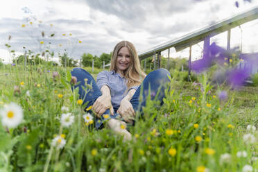 Happy engineer sitting on grass in front of solar panels at field - OSF00677