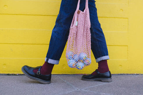 Junge Frau mit Globus in Netztasche geht an gelber Wand vorbei - ASGF02621