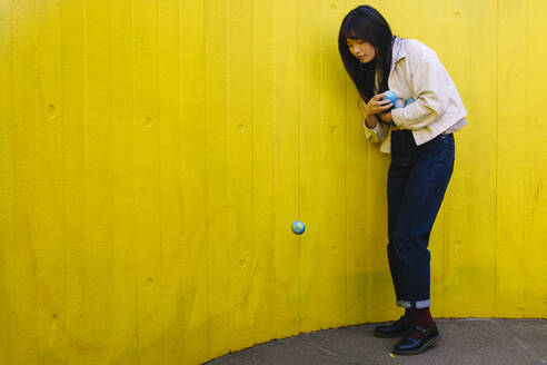 Young woman looking at fallen globe in front of yellow wall - ASGF02623