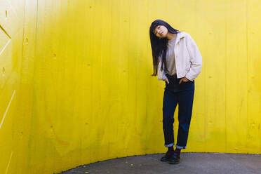 Junge Frau mit geneigtem Kopf vor einer gelben Wand stehend - ASGF02632