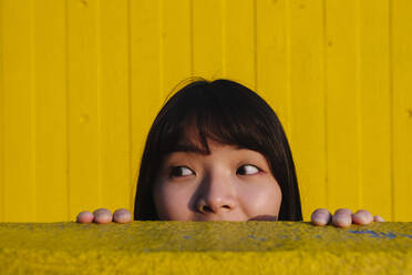 Young woman looking away peeking from wall - ASGF02647